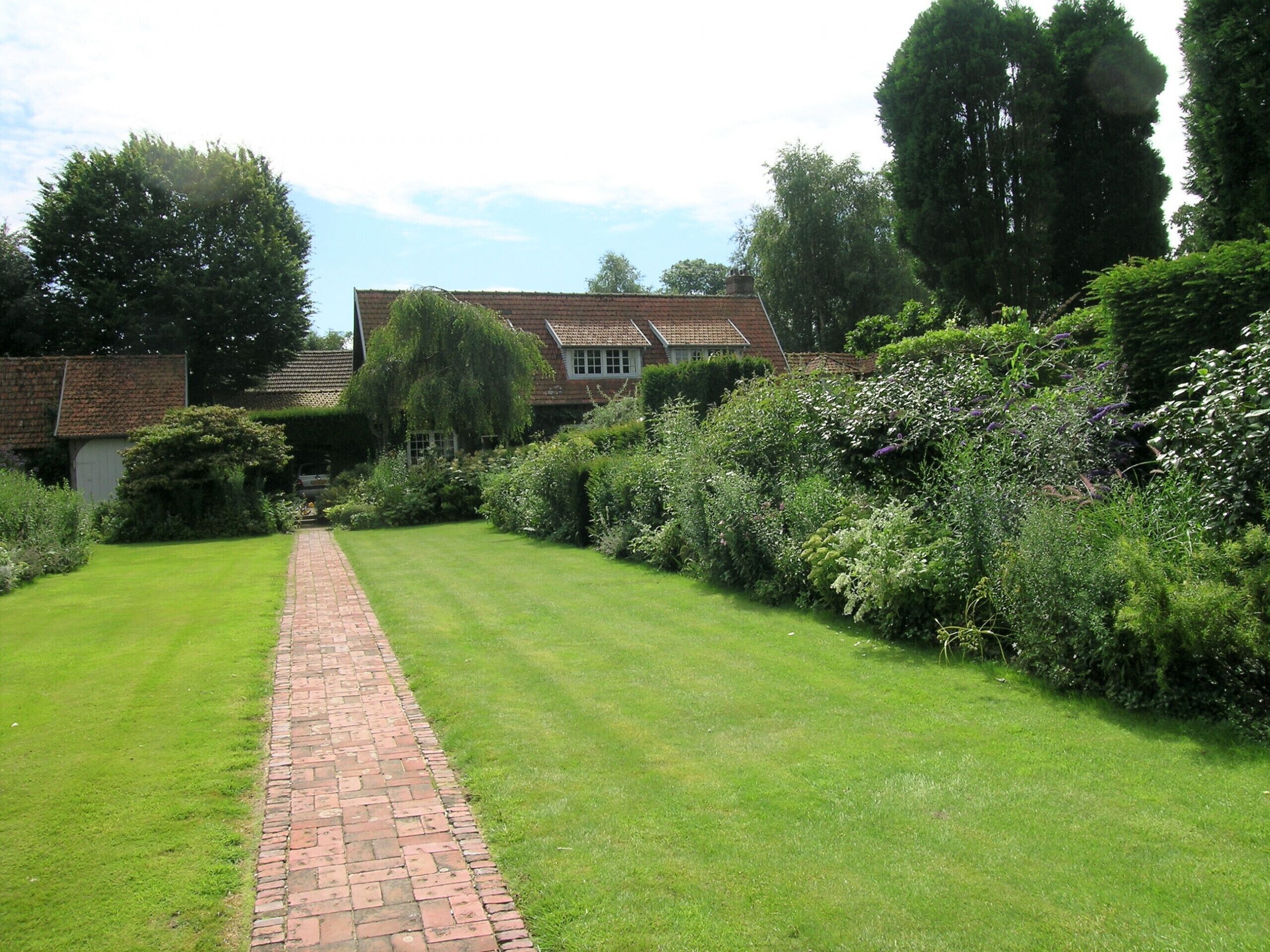 A brick pathway leading to a house with a tiled roof, surrounded by lush greenery and trees.