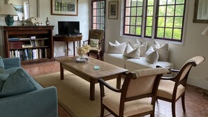 Uma sala de estar com sofá, poltronas, mesa de centro, estante de livros, televisão e janelas amplas com vista para o verde.