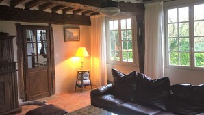 Ein gemütliches Wohnzimmer mit Ledersofa, Glastisch und Holzboden. Ein Fenster mit Vorhängen, eine Lampe auf einem Beistelltisch und ein gerahmtes Bild an der Wand sind zu sehen.