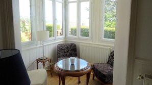 A corner room with a bay window, a round wooden table with a glass top, two patterned chairs, a floor lamp, and a radiator. - Consolata (Hautot-sur-Mer)