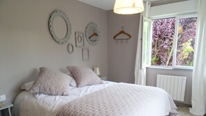 Et soveværelse med en stor seng, to puder, et natbord, en lampe, en radiator og et vindue med udsigt til træer.