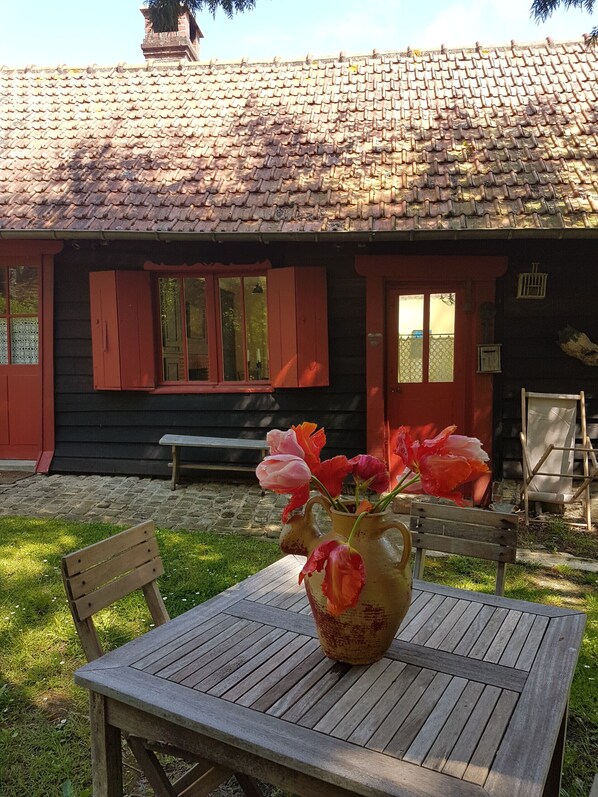 A rustic wooden house with a tiled roof and a wooden table with a vase of flowers in front.