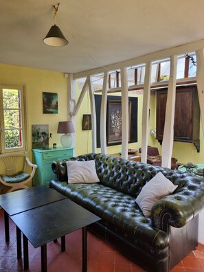 A cozy living room with a green leather sofa, a wooden chair, and a table. There are paintings on the walls and a window with a view of greenery.