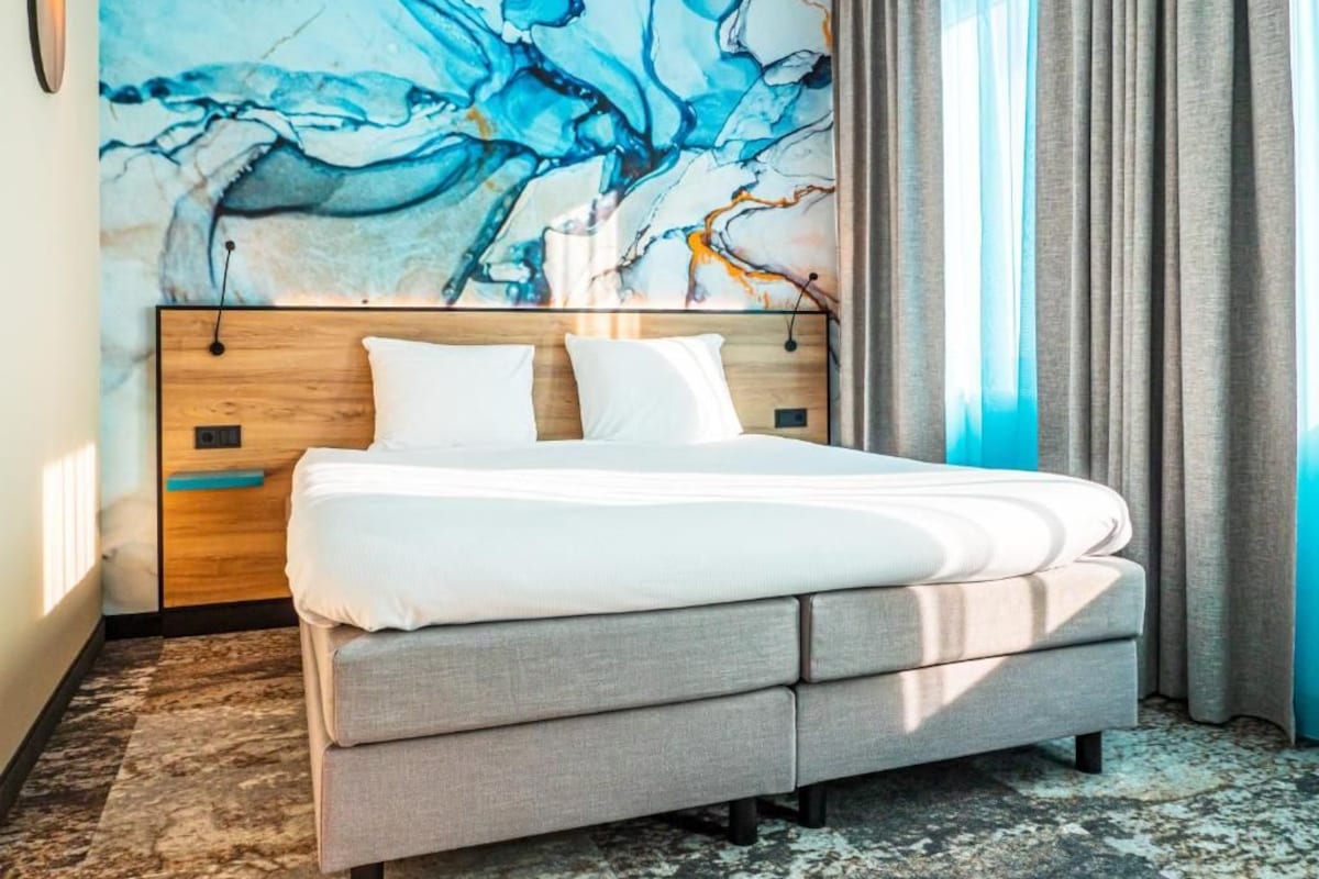A modern bedroom with a large abstract painting on the wall, a wooden headboard, and a grey upholstered bed.