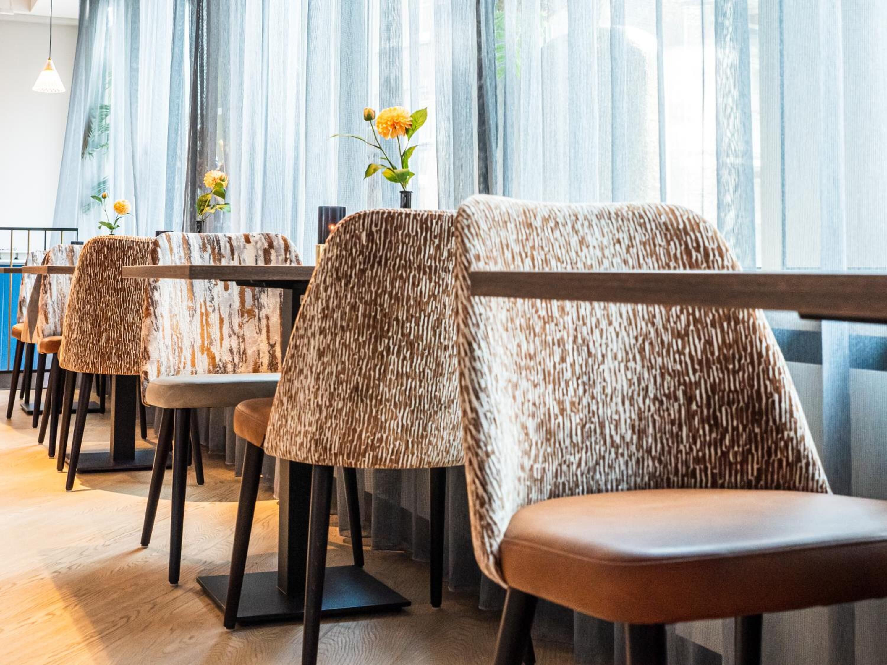 A restaurant with patterned chairs and tables, large windows with sheer curtains, and a wooden floor.