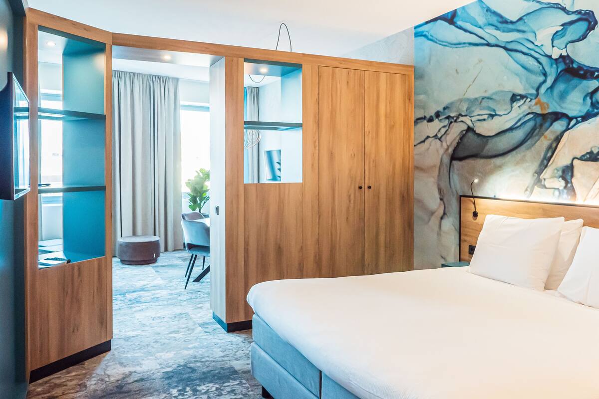 A hotel room with a large bed, wooden wardrobe, a desk with a chair, and a view of the outdoors.