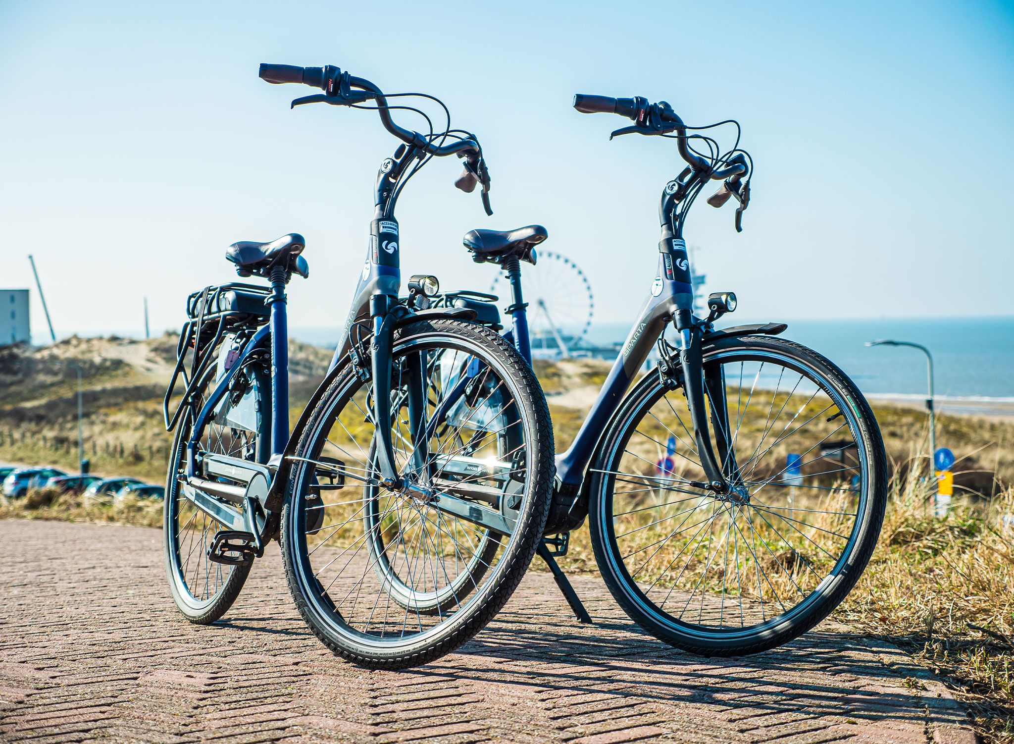 Dos bicicletas estacionadas en un camino pavimentado con vistas panorámicas al océano y una noria al fondo.