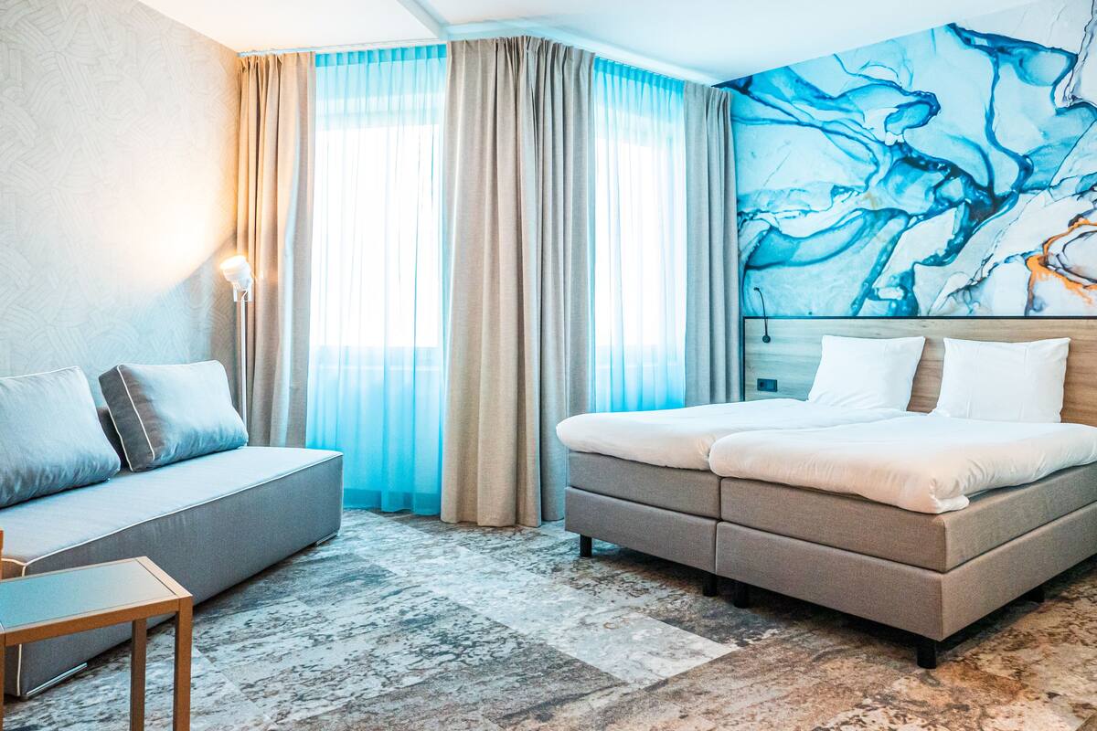 A hotel room with a bed, a sofa, a desk, and a large abstract painting on the wall.