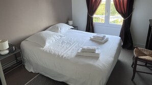 Una cama bien tendida con sábanas y toallas blancas, una lámpara de noche, una silla y una ventana con cortinas.