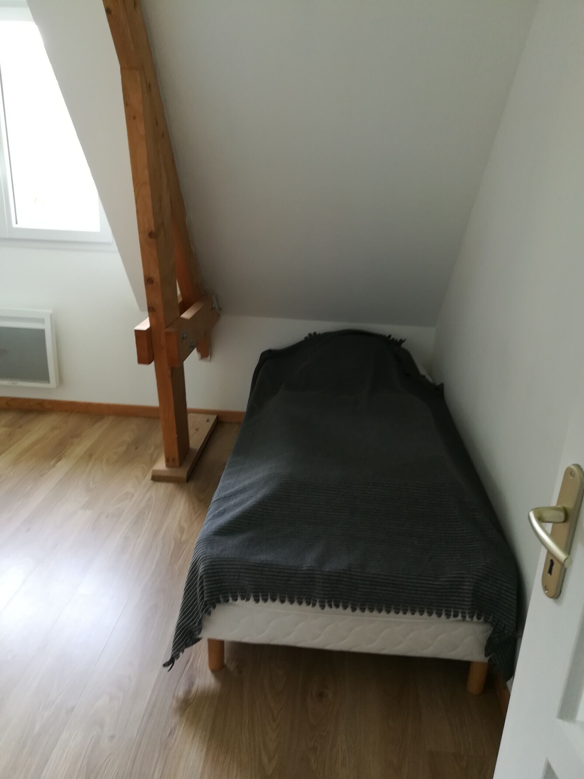 A small, narrow room with a wooden bed frame, a window, and a door with a brass handle.