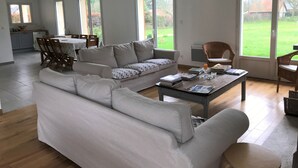 A spacious living room with a large sofa, a coffee table, and a dining area with chairs and a table. - Le Cèdre (Varengeville-sur-Mer)