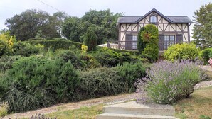 A traditional Tudor-style house with a symmetrical facade, a central tower, and a well-maintained garden with various shrubs and a gravel path leading to the entrance. - Valleuse (Saint-Léonard)