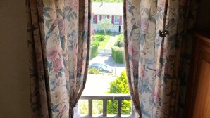 A window with floral curtains, a balcony, and a view of a house and greenery. - Valleuse (Saint-Léonard)