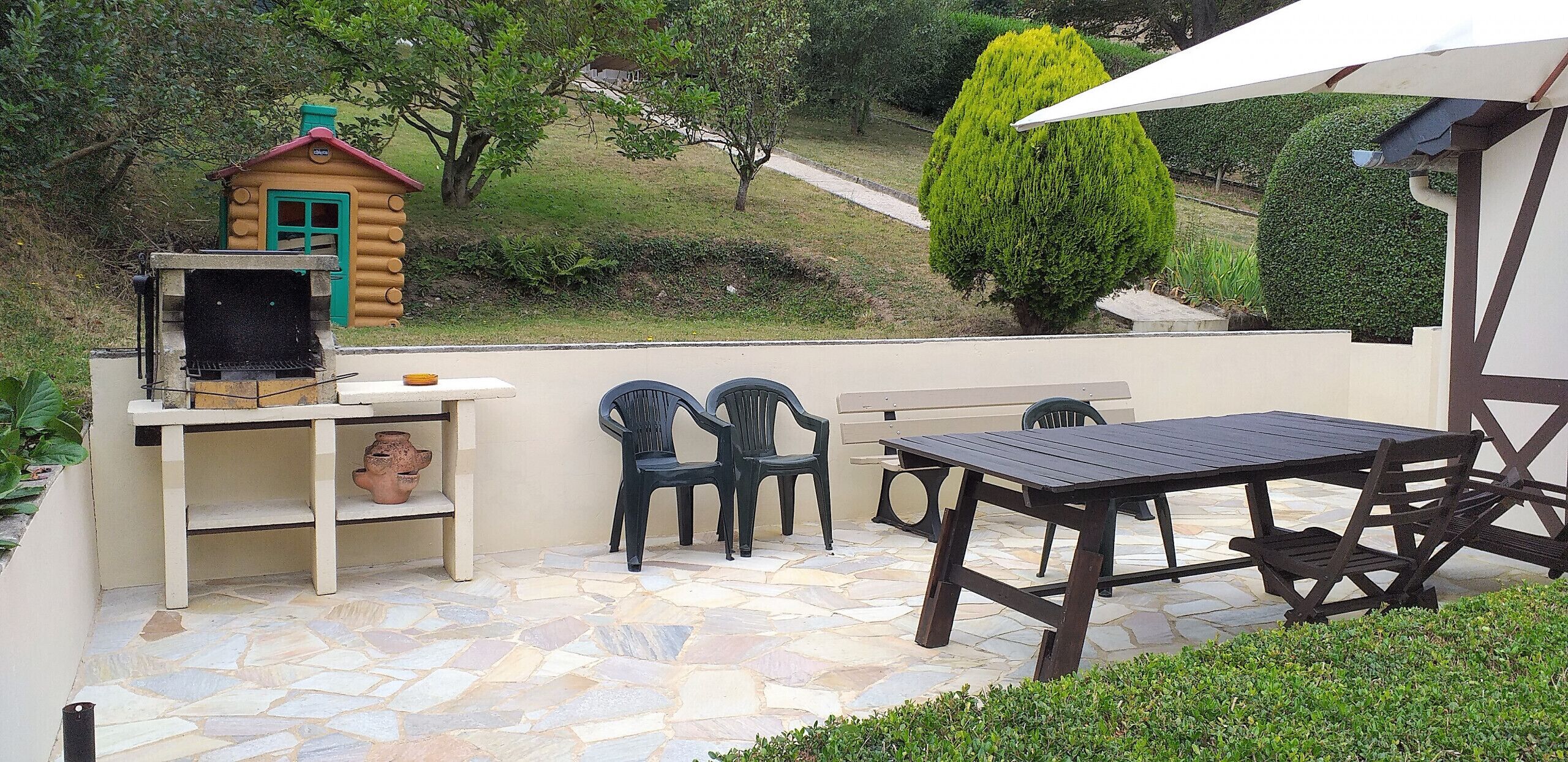 A patio with stone flooring, a wooden table, two plastic chairs, a small wooden structure with a chimney, and a green coniferous tree in the background.