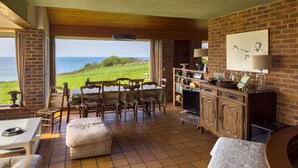A cozy room with a view of the ocean, featuring a wooden dining table, chairs, a sofa, and a TV.