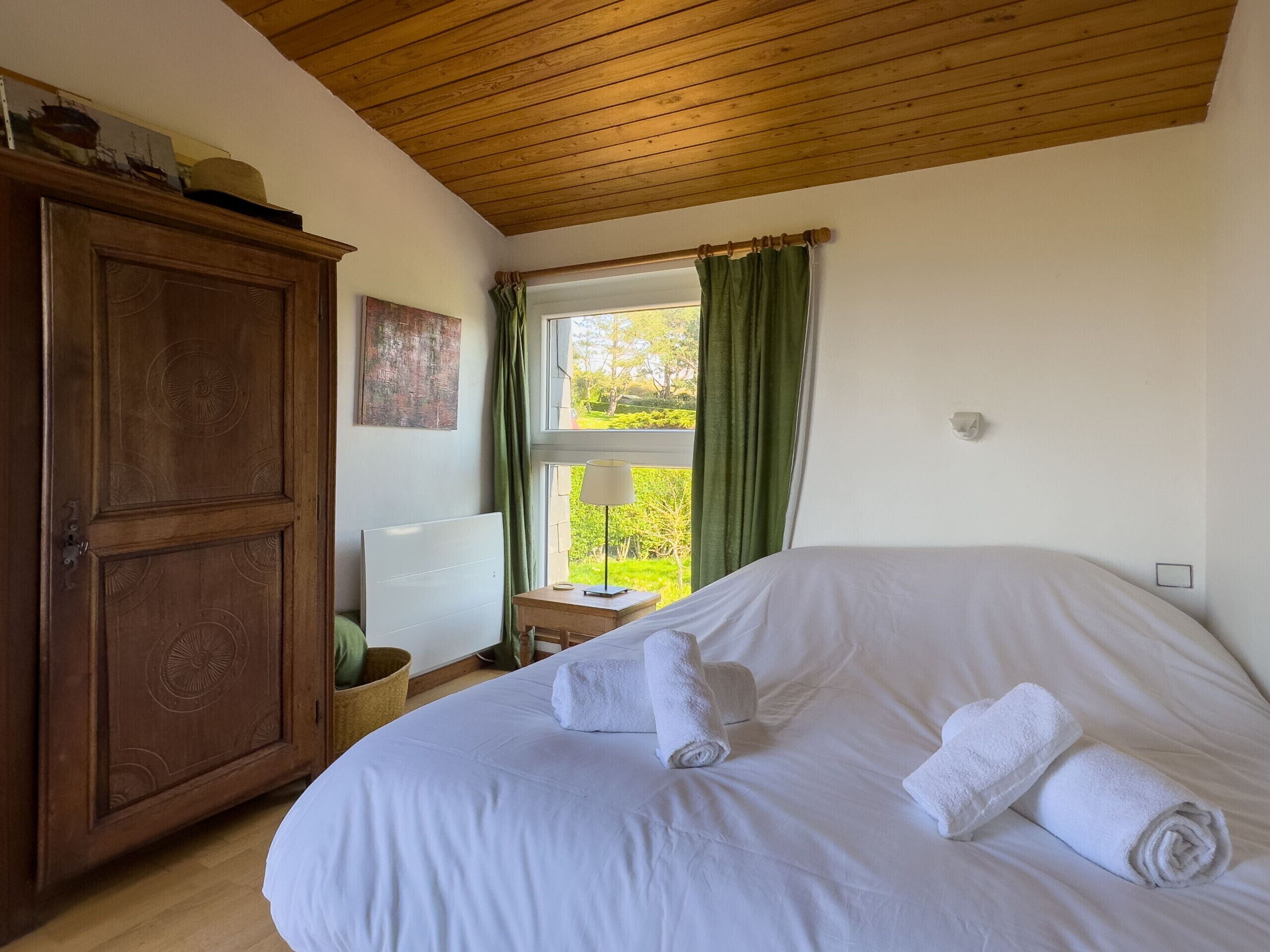 A bedroom with a large bed, a wooden wardrobe, a window with curtains, and a lamp on a bedside table.