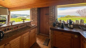 A rustic kitchen with wooden cabinets, a brick wall, and a large window offering a view of the countryside.
