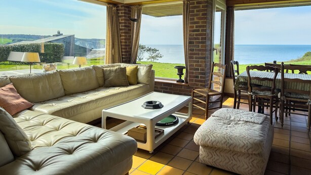 A cozy living room with a sofa, a coffee table, and a dining area with chairs and a table, all overlooking a scenic view of the sea.