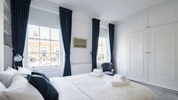 A neatly made bed with white linens and a grey chair, a white wardrobe, and a window with blue curtains.