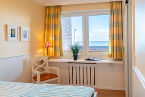 Quarto com cama, uma cadeira, janela com cortinas, radiador e quadros nas paredes.
