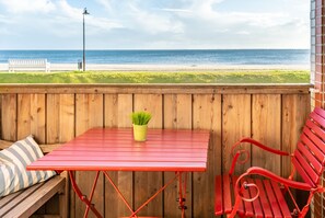Um deck de madeira com uma mesa e um banco de metal vermelho, com vista para uma praia.