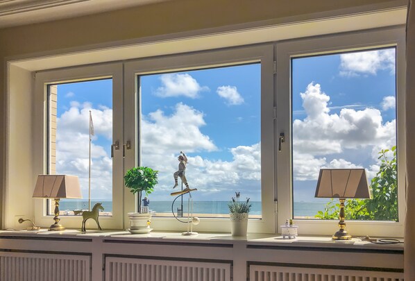 Quarto com três janelas grandes, cada uma com vista para o mar e para o céu. Há duas luminárias no parapeito da janela, uma pequena planta em vaso e uma estatueta de cavalo decorativa.