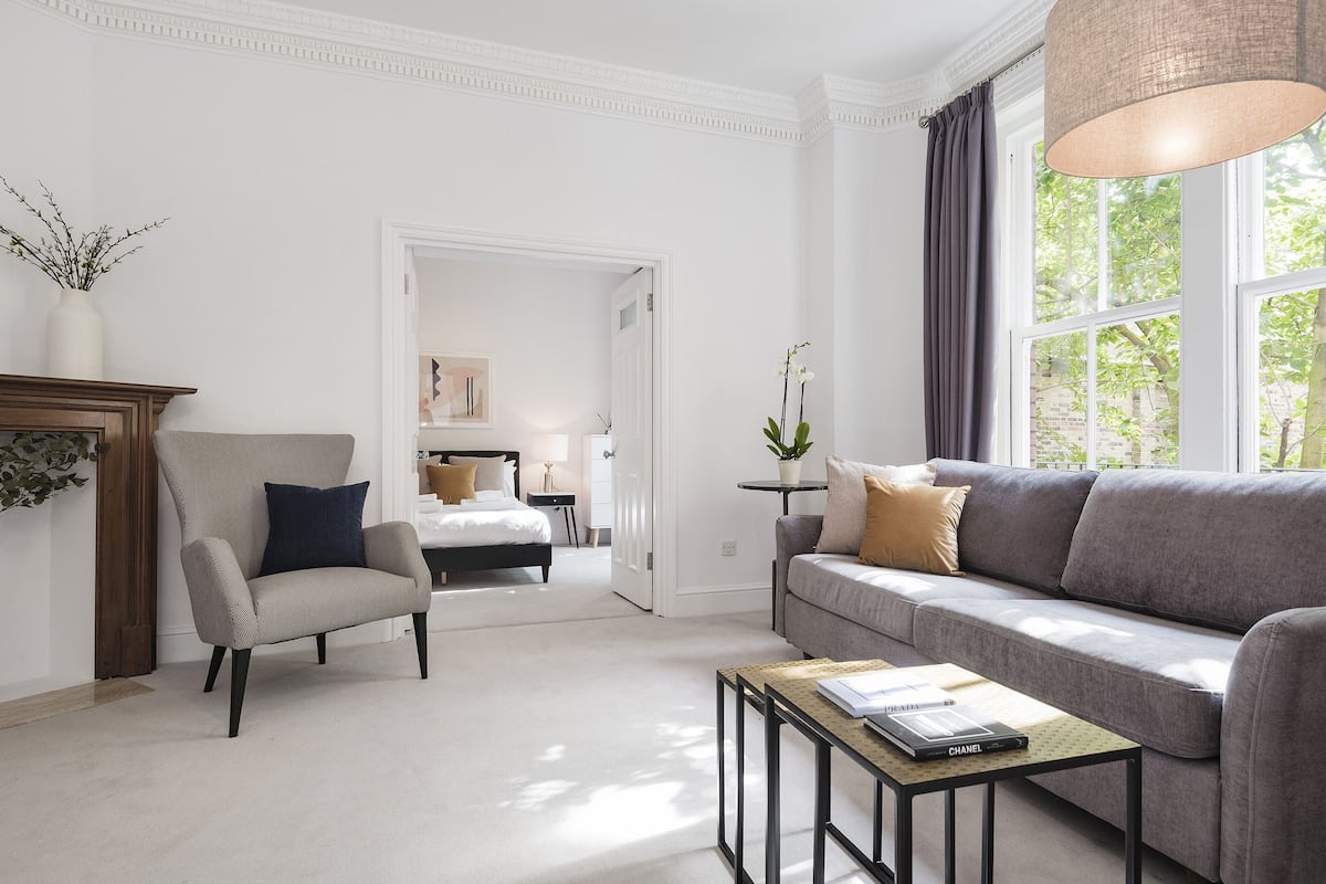 Un soggiorno moderno con un divano grigio, una poltrona abbinata e un tavolino da caffè. Un'apertura conduce a una camera da letto con un letto e un comodino.