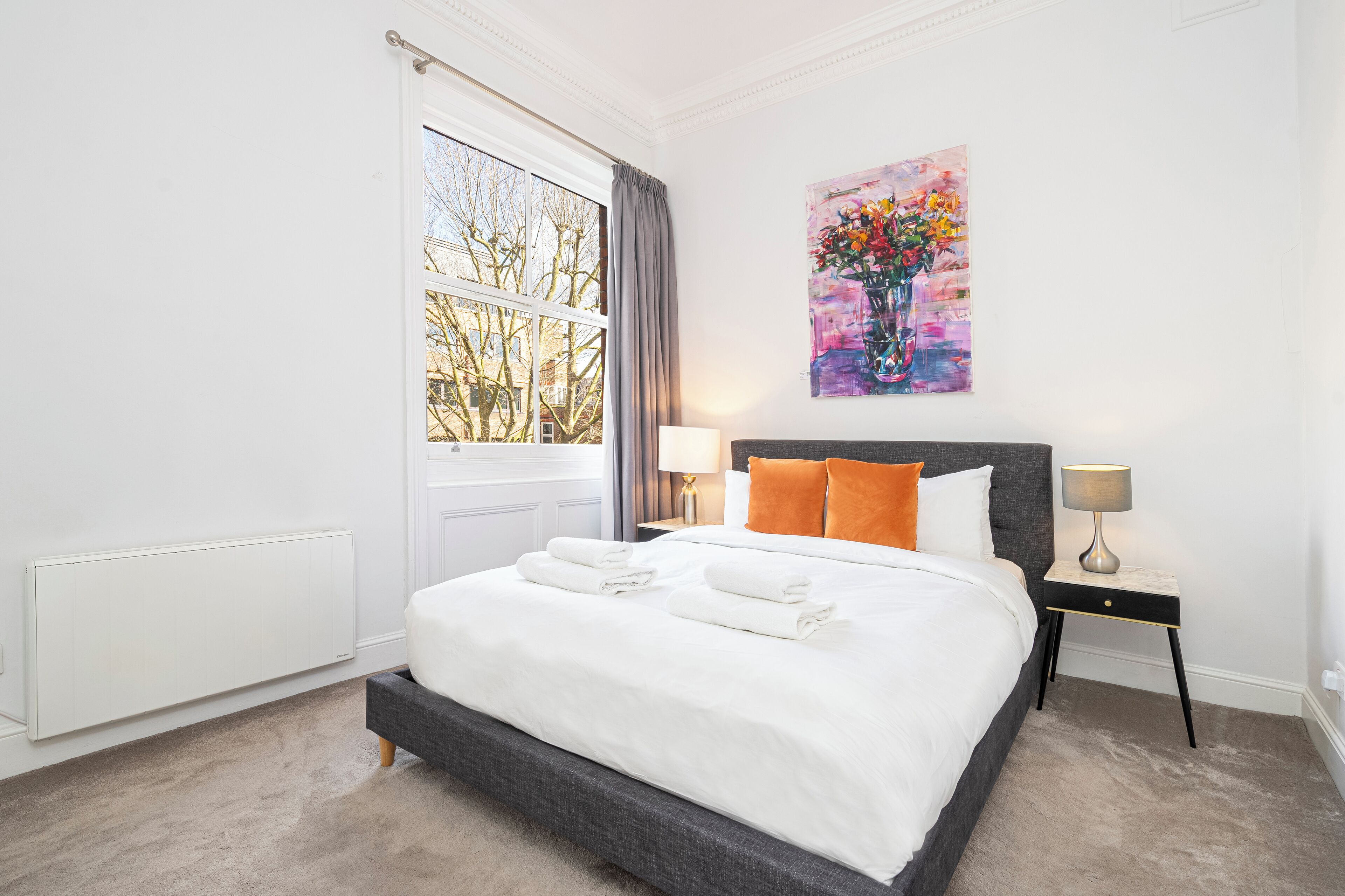 Un lit bien fait, avec du linge de lit blanc, une tête de lit grise et un oreiller orange. Un tableau représentant des fleurs est accroché au mur, il y a une fenêtre donnant sur des arbres et un radiateur blanc.