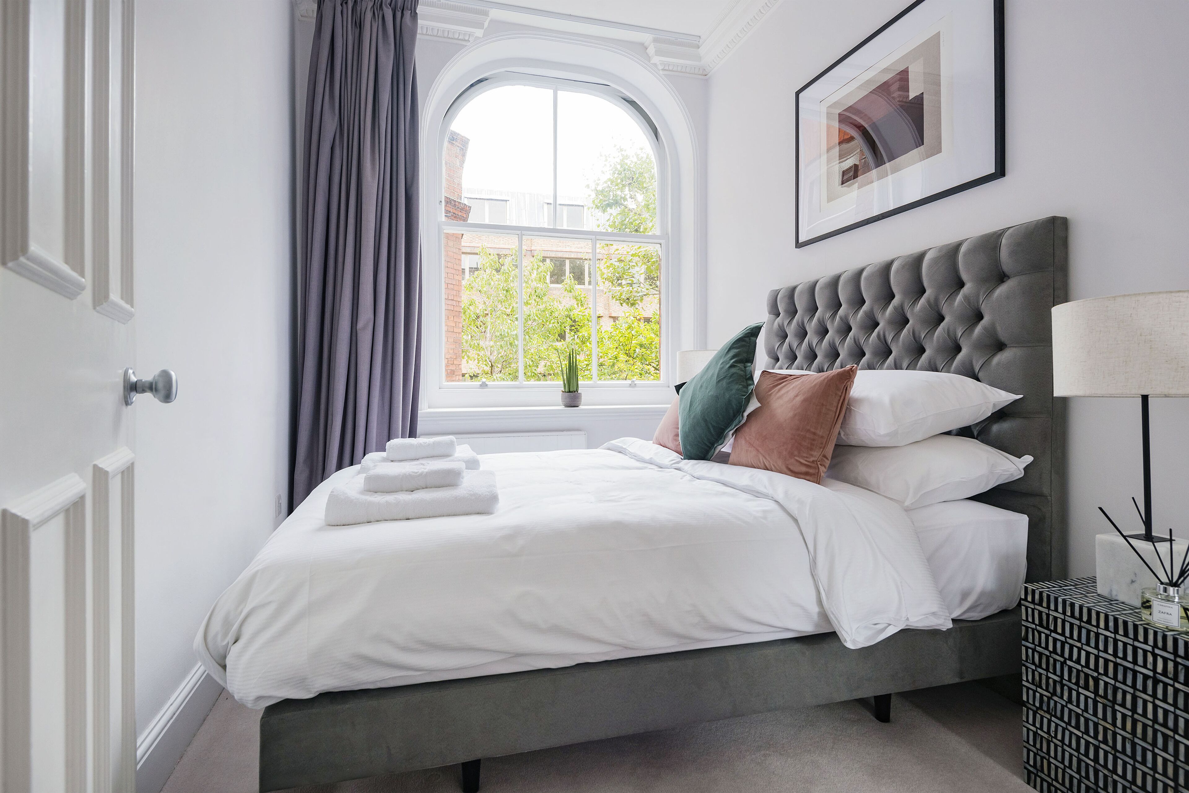 A neatly made bed with white linens and a grey headboard, a window with curtains, and a framed picture on the wall.