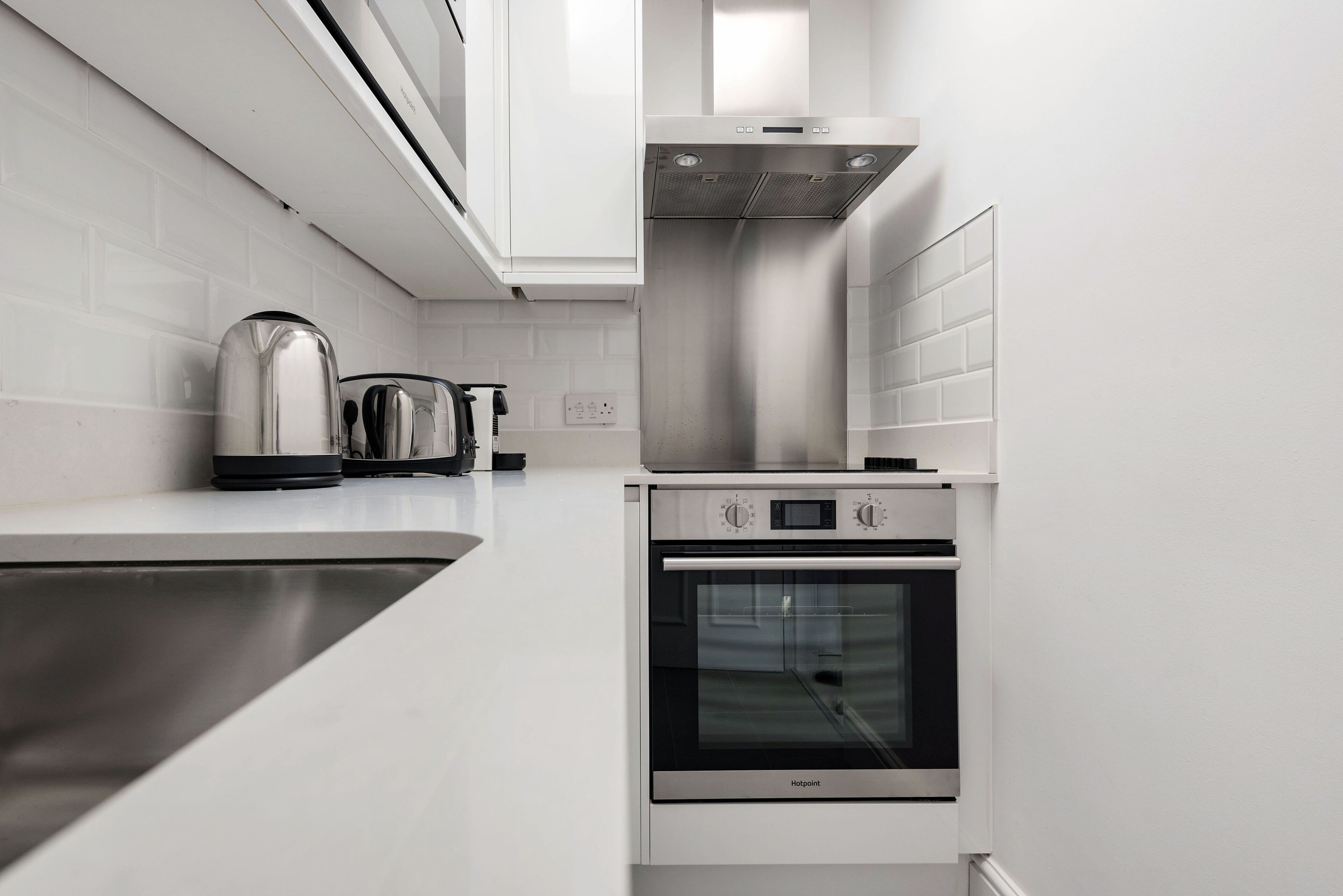 A compact kitchen with a stainless steel range hood, a built-in oven, and a kettle on the countertop.