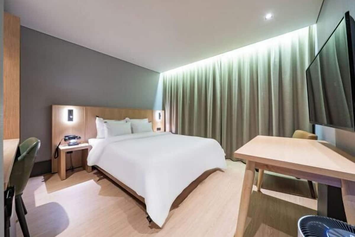 A modern hotel room with a bed, bedside table, chair, desk, and a flat-screen TV.