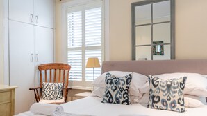 Un lit bien fait, agrémenté d’oreillers à motifs, une chaise en bois avec un coussin, une table de chevet et une fenêtre avec des stores.