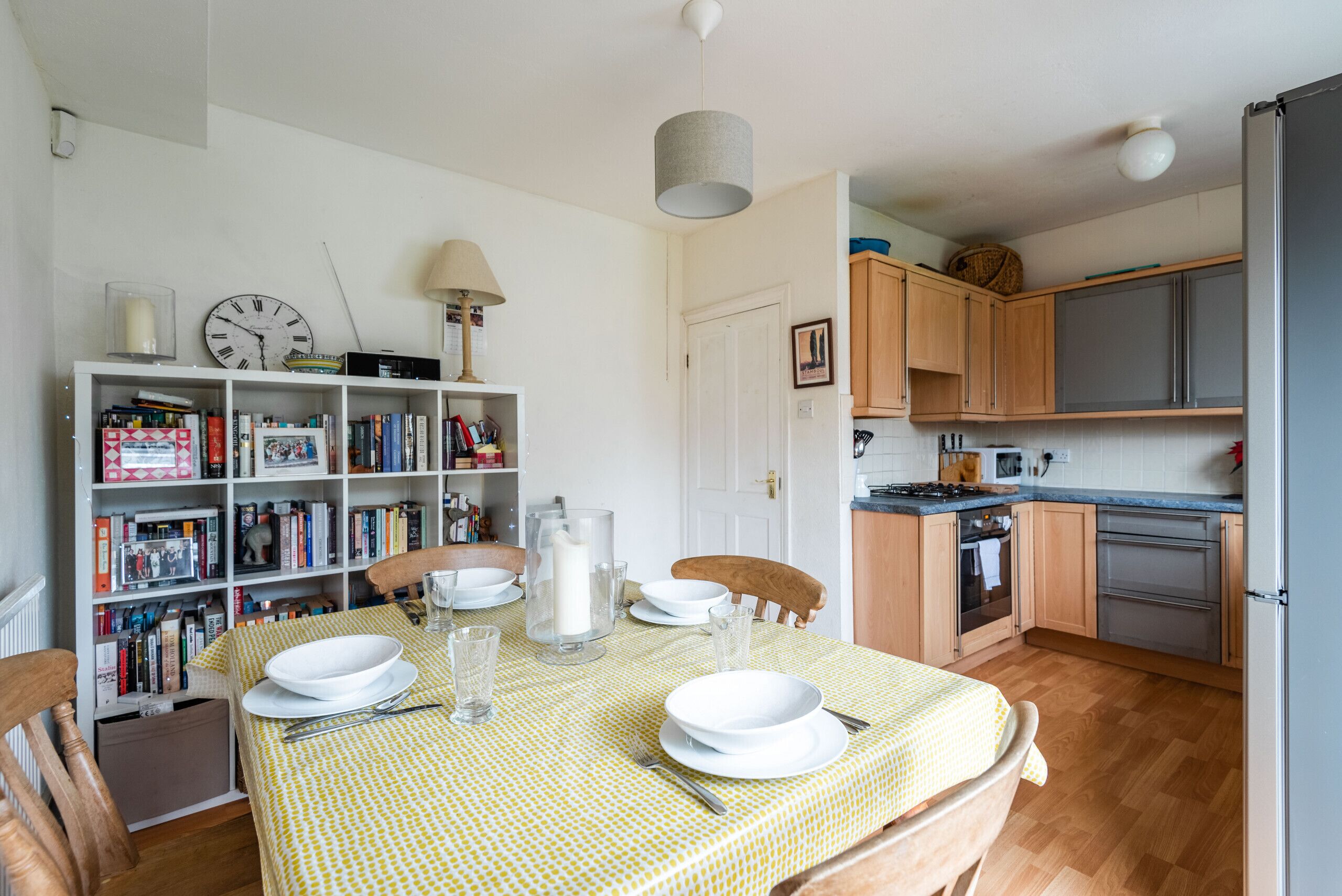 Un coin repas avec une table dressée pour quatre personnes, entourée de chaises en bois. Une bibliothèque remplie de livres et d’objets décoratifs se trouve à gauche. On aperçoit l’espace cuisine à l’arrière-plan, avec des placards en bois et un réfrigérateur.
