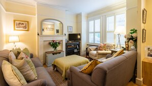 Un salon comprenant un canapé, un fauteuil et un téléviseur. On y trouve une cheminée avec une manteau de cheminée et un miroir au-dessus.