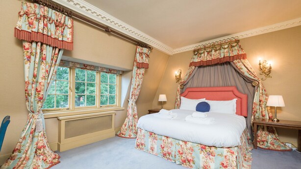 Une chambre à coucher avec un grand lit, des rideaux à motifs floraux, une fenêtre donnant sur des arbres et une table de chevet avec une lampe.