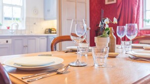 Une table à manger est dressée avec des assiettes, des couverts et des verres à vin dans une pièce bien éclairée, où l’on aperçoit un rideau rouge et une fenêtre en arrière-plan.