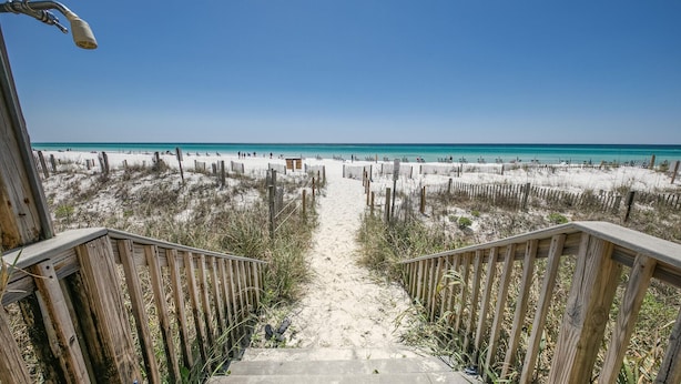 Un escalier en bois mène à une plage de sable fin, sous un ciel bleu et dégagé.