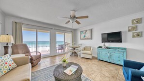 Un salon spacieux avec vue sur la plage, doté d’un ventilateur de plafond, d’une télévision et d’un meuble bleu.