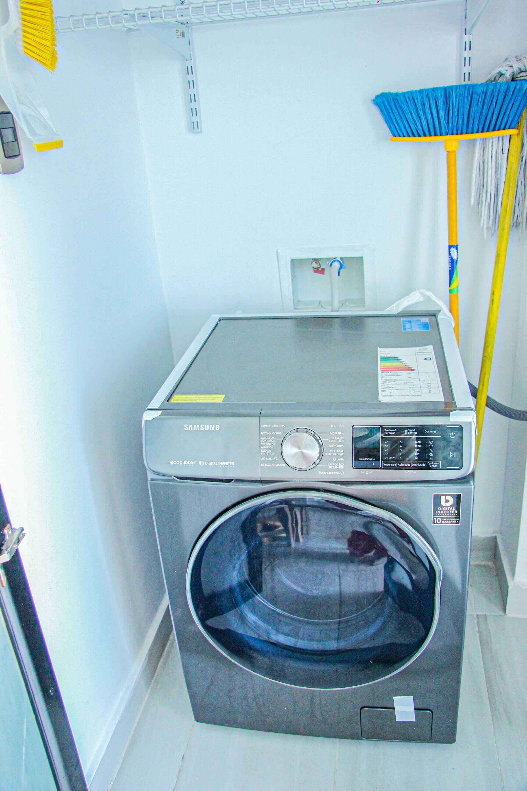 A Samsung digital inverter washing machine in a laundry room with a broom and a shelf above.
