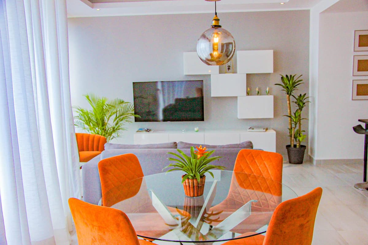 A modern living room with a glass dining table, orange chairs, a flat-screen TV, white shelving, and potted plants.
