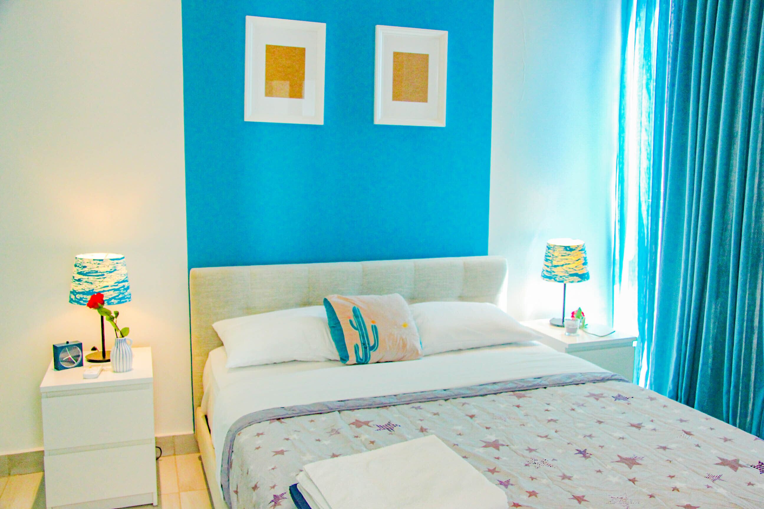 Un dormitorio con cama, dos mesitas de noche, una lámpara, un jarrón con una flor y una ventana con cortinas.