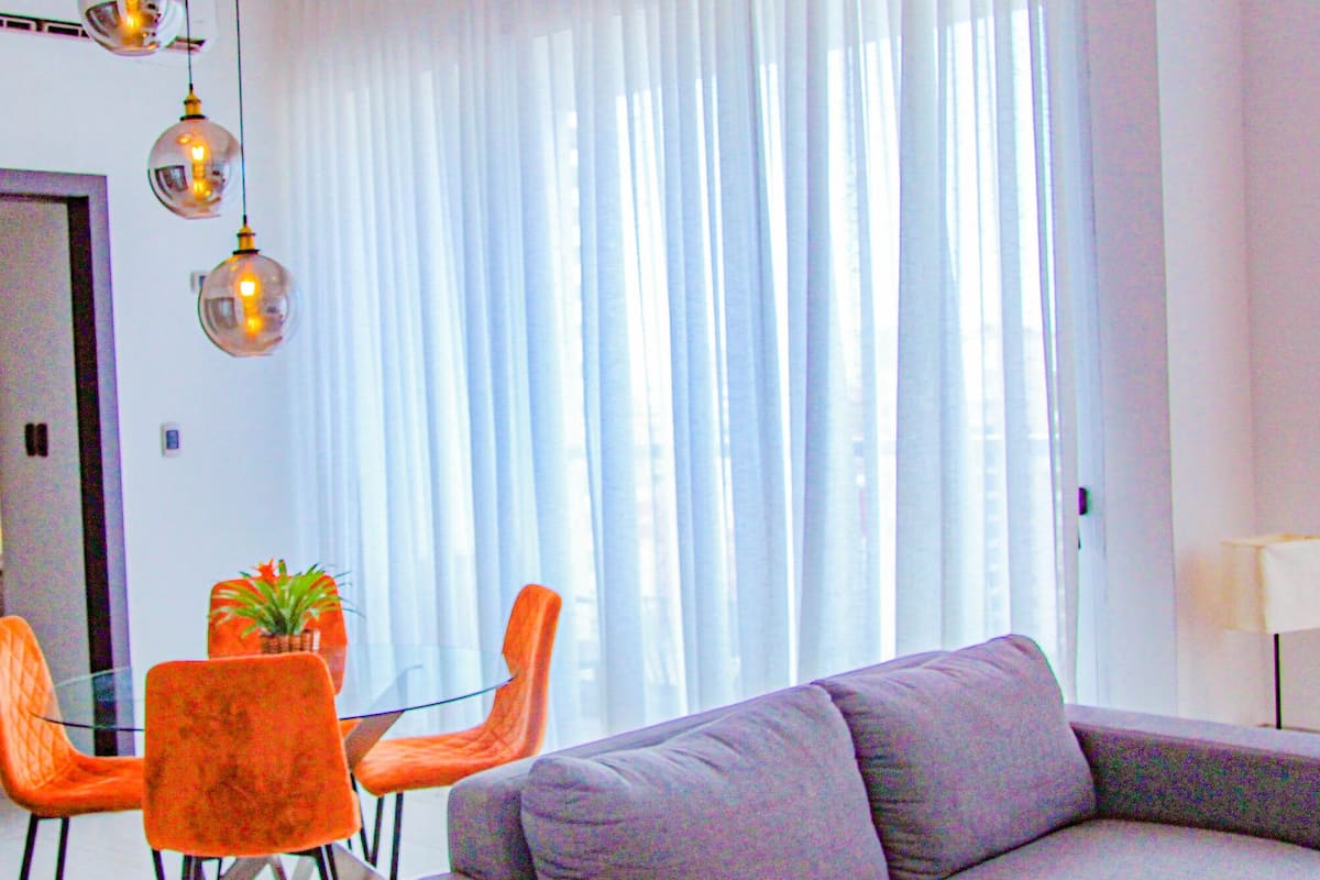 A modern living room with a grey sofa, orange chairs, a glass table, and a potted plant.