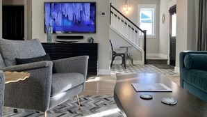 Una sala de estar con un sofá gris, una mesa de centro, un televisor en la pared y una escalera que lleva a otra habitación.