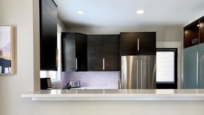 Una cocina moderna con armarios oscuros, electrodomésticos de acero inoxidable y una encimera blanca. Hay tres taburetes de madera en la barra.