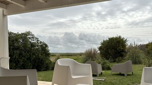 An outdoor patio with white plastic furniture, a tiled floor, and a view of a grassy landscape with trees and a cloudy sky.
