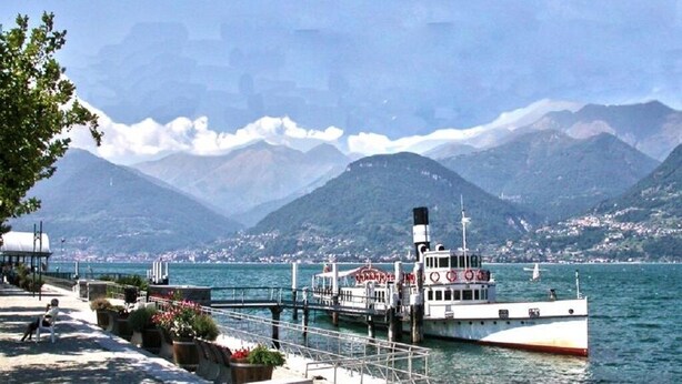 A waterfront promenade with a boat docked, mountains in the background, and a clear sky.
