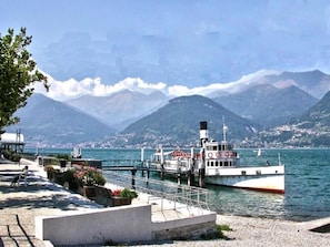 A waterfront promenade with a boat docked, mountains in the background, and a clear sky.