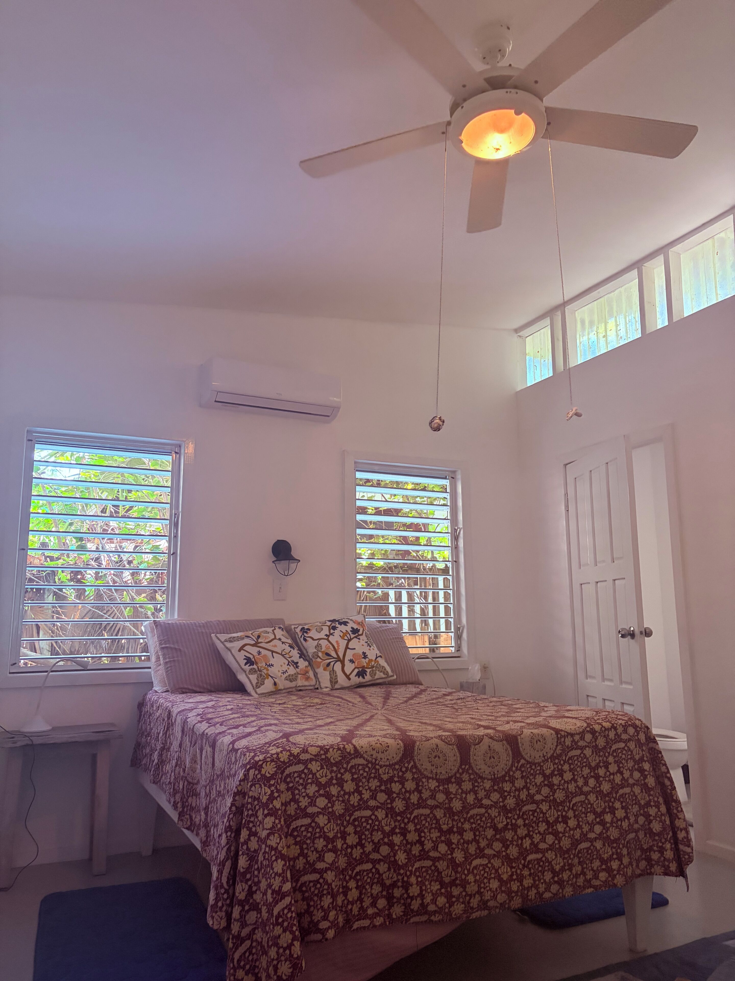 Ein Schlafzimmer mit einem Bett, zwei Fenstern, einem Deckenventilator und einer Klimaanlage.