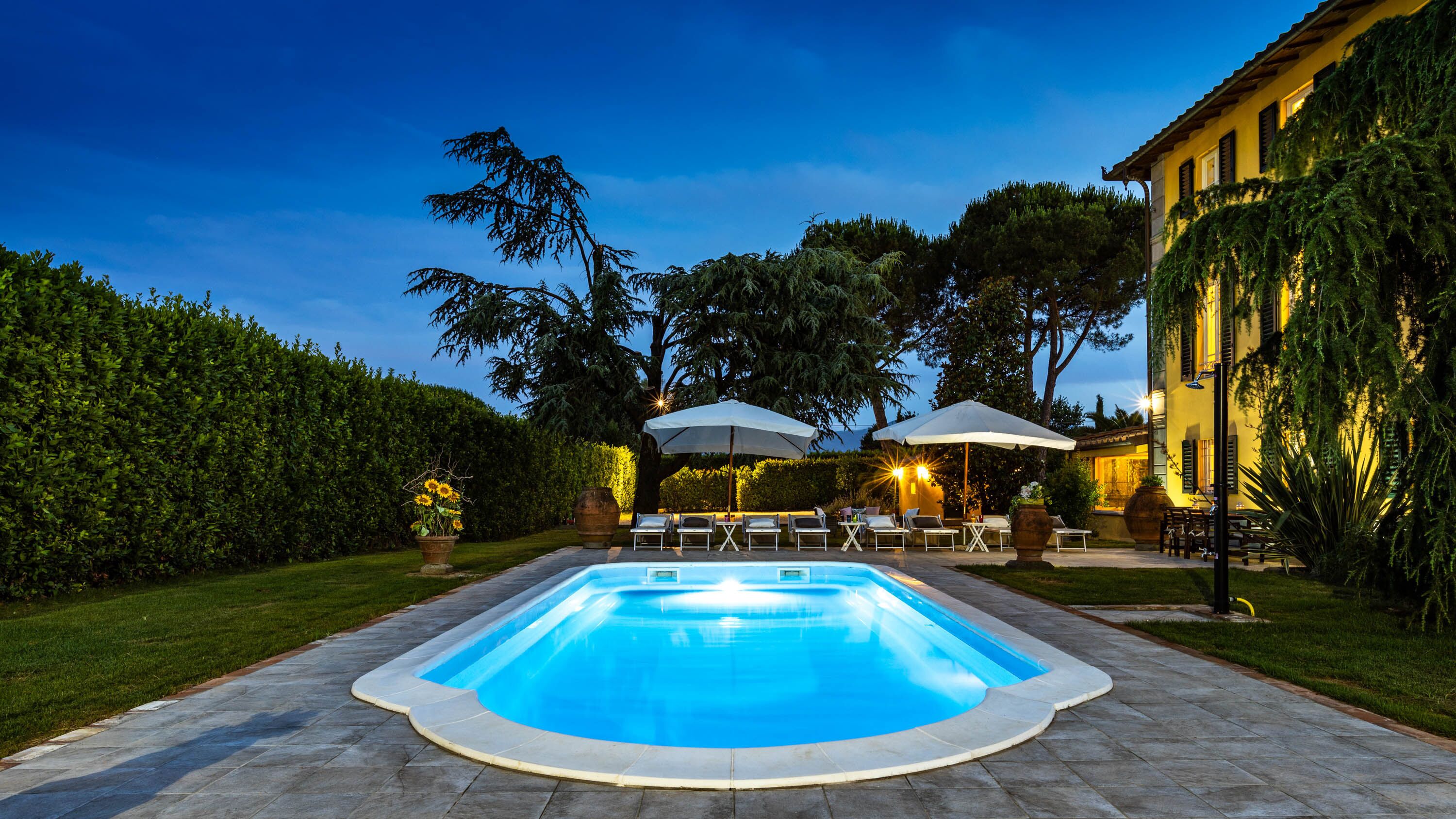 A rectangular swimming pool with a tiled deck, surrounded by a well-maintained hedge and trees. There are lounge chairs and umbrellas under a pergola, and a building with yellow walls and shutters in the background.
