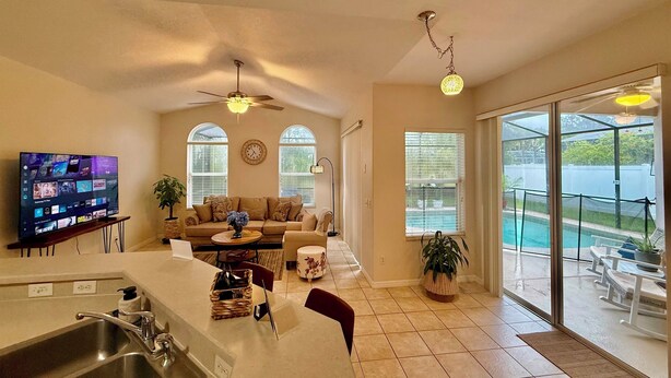 A spacious living area with a large TV, a ceiling fan, and a view of a swimming pool. - Grand Village 4BR Pool Home near Disney World (Davenport)